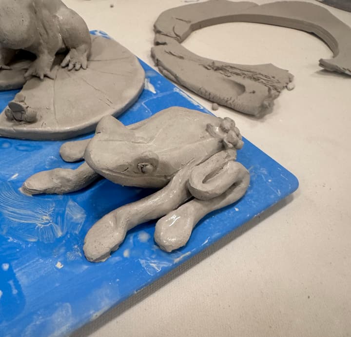 Hand-sculpted clay frog on a blue work board beside a frog on a lily pad