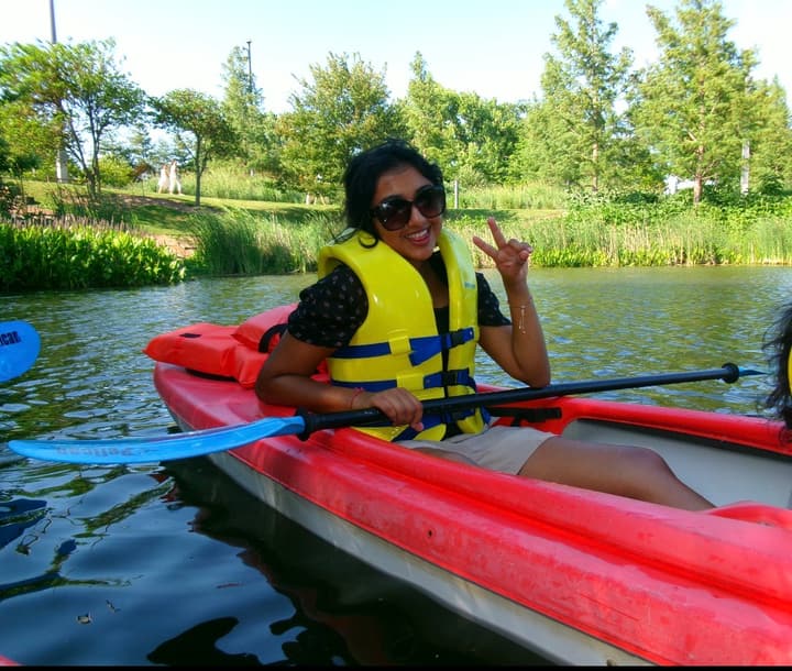 Smiling in a yellow life jacket and sunglasses in a red kayak on a tree-lined lake