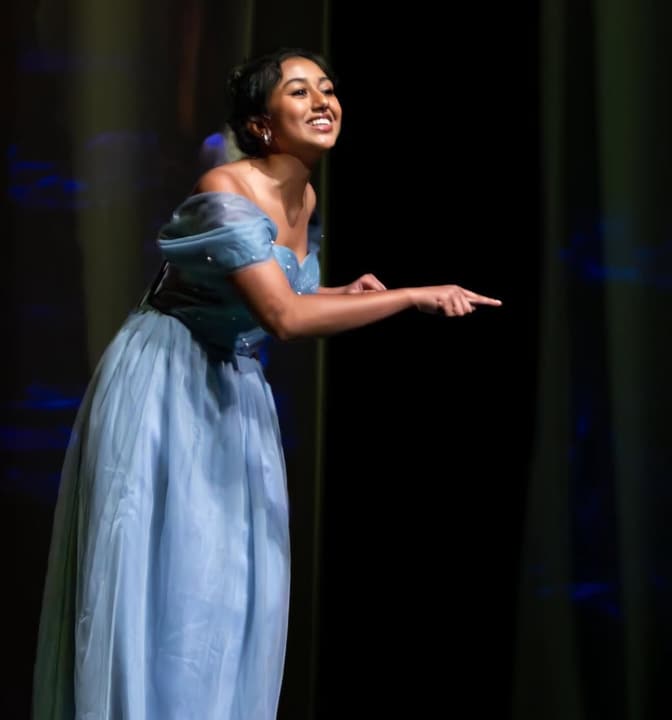 Performer in a pale blue gown leaning forward and pointing on a dark stage
