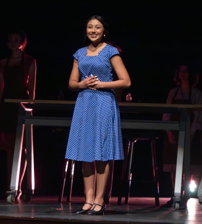 Performer in a blue polka-dot dress standing center stage under a spotlight