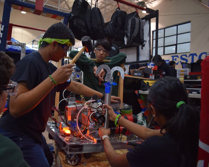 Robotics teammates in safety glasses assembling a competition robot in a gym pit area, one swinging a mallet